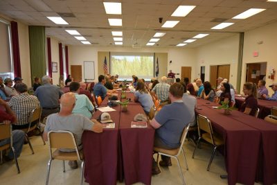 A group seated at tables views a video on a large screen.