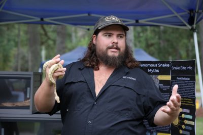 A man with a beard wearing a hat holds a snake in his hand while speaking at Family & Farm Day.