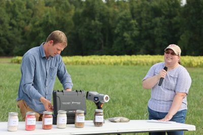 A man behind a table prepares a hand-held device for use as a woman with microphone speaks to the field day attendees.