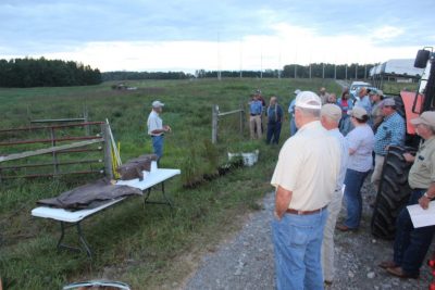 A man in cap stands beside a table as he speaks to the field day attendees lining the edge of a field.