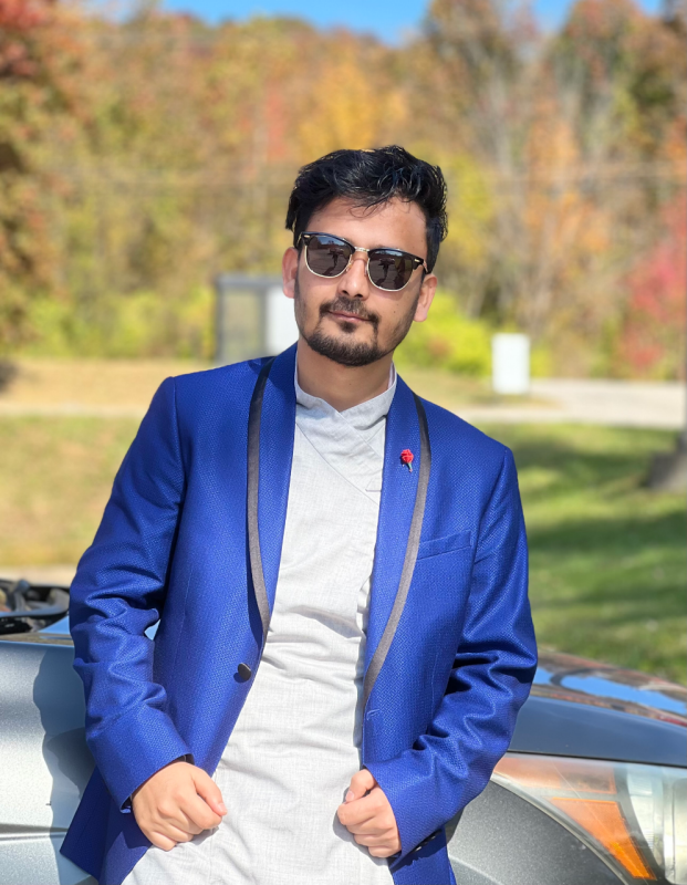 Young man wearing blue jacket and sunglasses leaning against car.