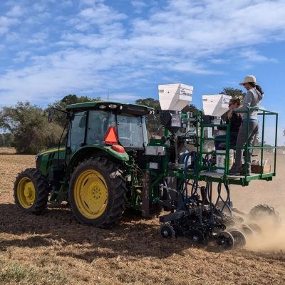 Two women ride on a planter behind a John Deere tractor in the field.