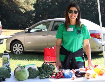 A woman in sunglasses and green shirt smiles from behind a table filled with different fruits and vegetables